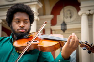 In front of Greek columns and sporting a green shirt, a mustache, beard, and mid-length natural African hair, Wilfred Farquharson plays his viola while looking towards the instrument
