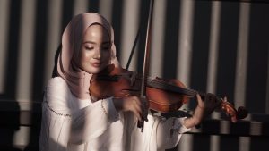  in front of a wall dappled with light, a light-skinned woman wears a coral-colored hijab, face makeup, and a white long-sleeved outfit while bowing her violin near the frog