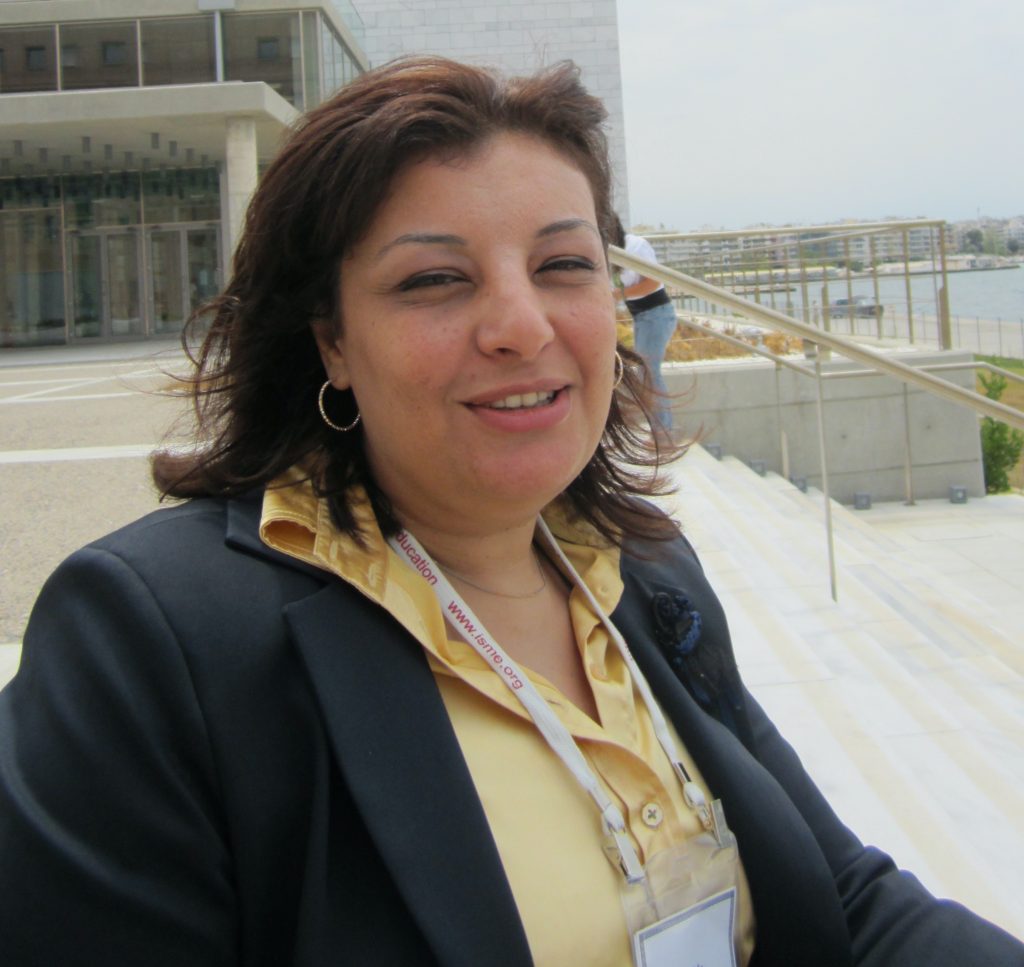 Composer Nahla Mattar is standing on the steps in front of a building. Nahla is wearing a dark blazer and yellow shirt, and her eyes are squinting as one does in bright sunlight.