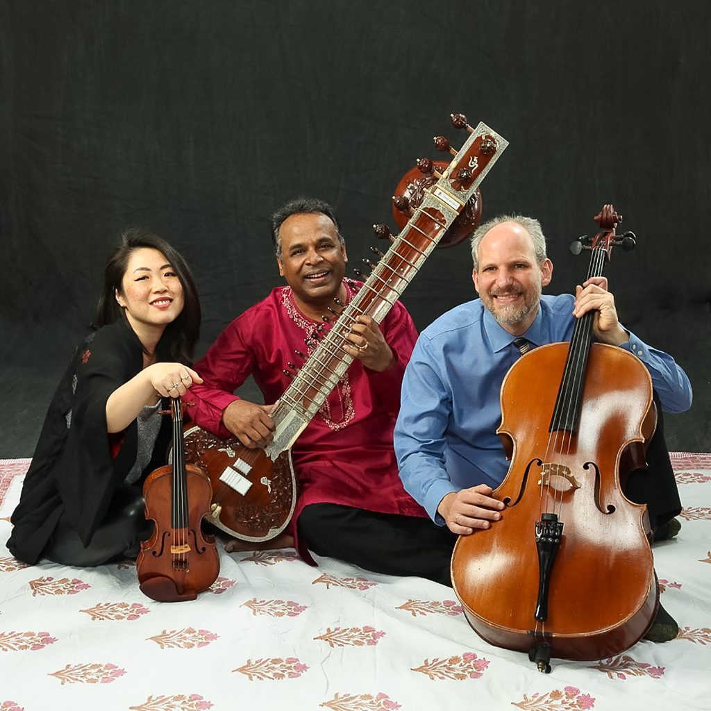 Violinist Maya, Sitarist Guarav, and Cellist Tom seated holding their instruments, smiling