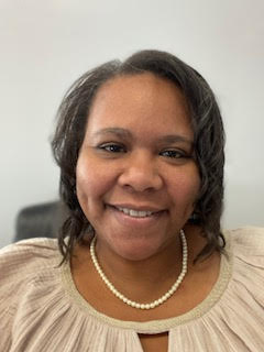 Before a white background, Dr. Casandra Holliday, a dark-skinned woman wearing a dressy white shirt and a pearl necklace, smiles at the camera