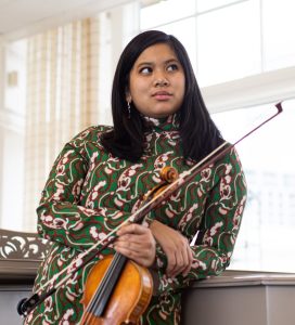 Wearing a green and pink patterned dress, Caroline Jesalva, an Asian woman, holds her violin and bow and leans against a grey piano