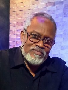 In front of a light purple brick background, Haitian composer Dickens Princivil wears a black collared shirt and glasses and sports a salt-and-pepper goatee and beard and glasses while looking towards the camera earnestly