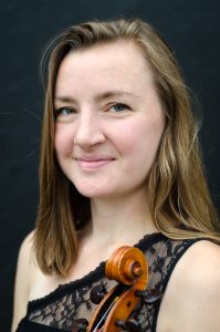Wearing a lacy black top, Hanna Pederson, a light-skinned woman with long straight blond hair, looks towards the camera and smiles while holding her viola