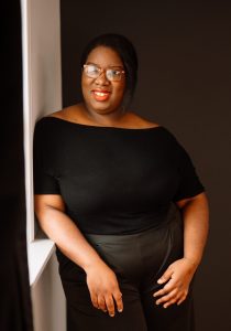 Standing in front of a dark wall, leaning on a wall to her side painted white near a window from which gentle light streams in, Composer Jessica T Carter, a dark-skinned woman sporting a black top and black pants, glasses, red lipstick, and straight, shoulder-length black hair, poses with poise and smiles gently towards the camera
